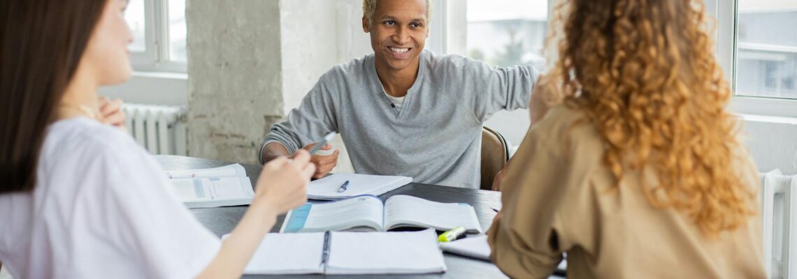 Positive African American male student and unrecognizable classmates looking at each other while sitting at table with textbooks and copybooks during lesson in classroom