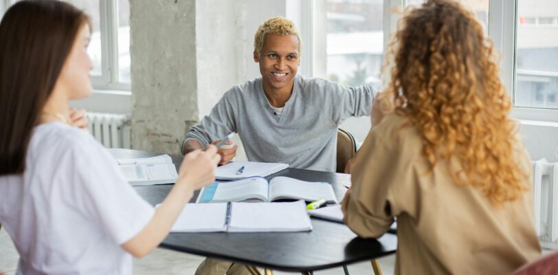 Positive African American male student and unrecognizable classmates looking at each other while sitting at table with textbooks and copybooks during lesson in classroom